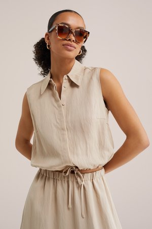 Žena s kučeravými vlasmi v copu, ktorá nosí veľké slnečné okuliare s rámom v tvare korytnačky, zlaté kruhové náušnice a bezrukávový béžový top na gombíky so zodpovedajúcimi nohavicami so šnúrkami.