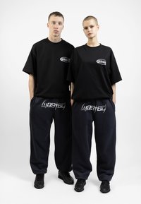 Camisetas negras oversized con logo en el pecho, combinadas con pantalones de chándal azul marino con texto gráfico blanco; ambos llevan zapatillas negras.