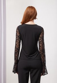 Un haut noir avec de longues manches en dentelle transparente et des poignets évasés. Corps en tissu lisse, encolure ronde et design ajusté. Vue arrière.