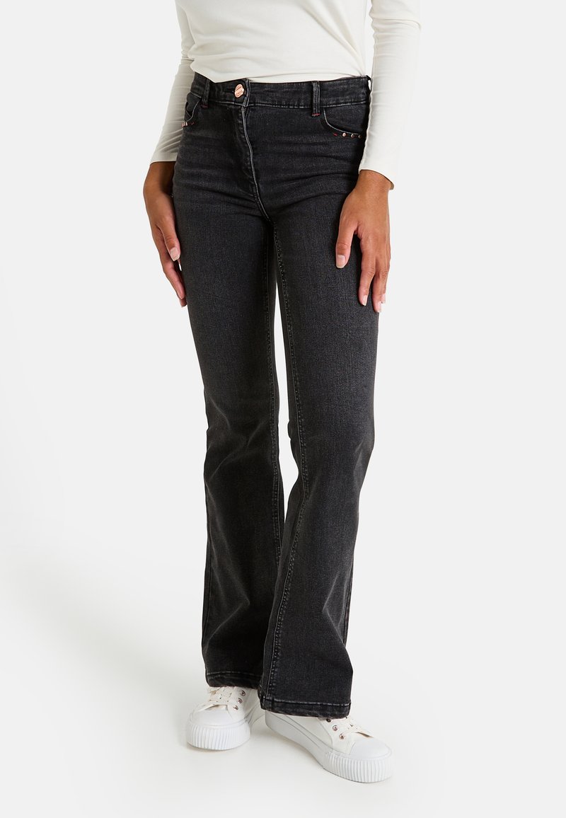 Mujer vestida con jeans de bota ancha negros, camiseta de manga larga blanca y zapatillas blancas, de pie con las manos relajadas a los lados.