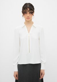 Camicia bianca a maniche lunghe con scollatura con zip, texture liscia e colletto strutturato, abbinata a una gonna scura a righe sottili.