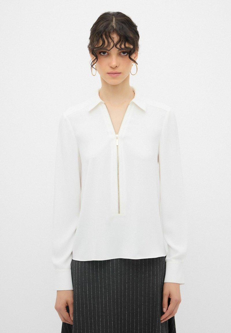 Camicia bianca a maniche lunghe con scollatura con zip, texture liscia e colletto strutturato, abbinata a una gonna scura a righe sottili.