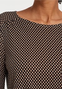 Svart topp med en avslappnad halsringning och ett mönster av små krämfärgade fyrkanter över hela. Tyget verkar slätt med en lätt textur.