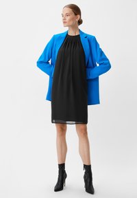 Czarna sukienka bez rękawów z marszczoną linią dekoltu, nałożona pod jasnoniebieską marynarkę. Modelka nosi czarne botki za kostkę z zadziornym noskiem.