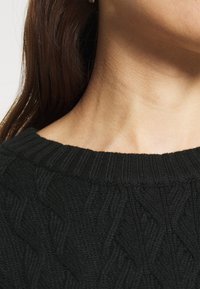 Pull en maille noire avec un col côtelé et un motif torsadé ; tissu texturé visible autour du col et de la zone des épaules.