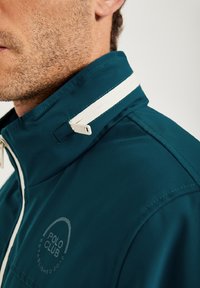 Tealfärgad jacka med hög krage, vit dragkedja och logotyp. Slät textur med en subtil lyster och sydda detaljer.