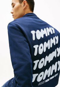 Marineblauwe jas met een gestructureerde afwerking. Bevat witte opbollende tekst "TOMMY" herhaald op de achterkant en "JEANS NEW YORK CITY" aan de onderkant.