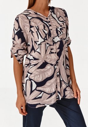 Mujer llevando una camisa holgada abotonada con grandes patrones de hojas beige y azul marino y mangas arremangadas, combinada con pantalones oscuros.