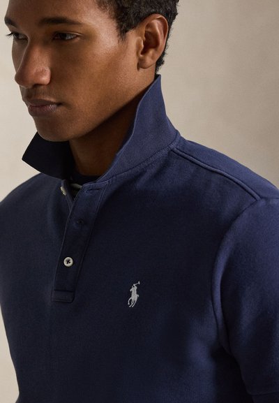 Άνδρας που φοράει μπλε ναυτικό πόλο πουκάμισο με όρθιο γιακά, με λογότυπο λευκού κεντημένου αλόγου και αναβάτη στο στήθος.