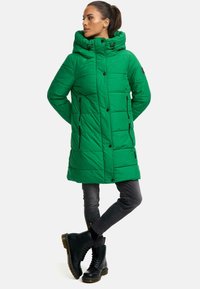 Abrigo acolchado verde con capucha, que presenta un diseño acolchado, botones frontales y bolsillos con cremallera negros. Lleva puesto sobre pantalones gris y botas negras.