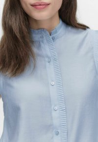 Lys blå, kortærmet bluse med stående krave, der har en knap-lukning fortil og lodrette flæse detaljer langs placket.