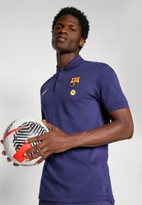 Navy polo shirt with FC Barcelona logo and Spotify emblem. The figure holds a black-and-white patterned soccer ball with a red accent.