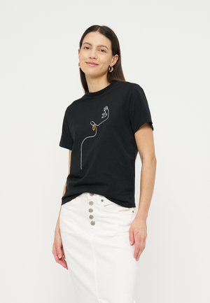 Γυναίκα με μακριά σκούρα μαλλιά φοράει μαύρο μπλουζάκι με αφηρημένη γραμμή προσώπου και χρυσούς κρίκους στα σκουλαρίκια, συνδυασμένο με λευκή φούστα με κουμπιά.