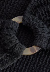 Svart texturerad scrunchie med en oval genomskinlig plastring. Tyget visar intrikata samlade mönster som ger djup åt designen.