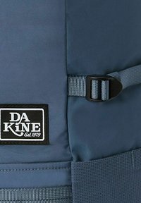 Dark blue backpack with a smooth, matte surface, featuring a black DA KINE logo patch and a plastic buckle strap. Textured mesh accents.