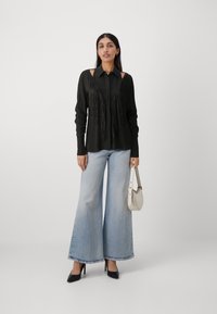 Svart skjorte med frynse detaljer, lange ermer og utskårne skuldre. Kombinert med lyseblå vide jeans og svarte hæler. Holder en hvit veske.