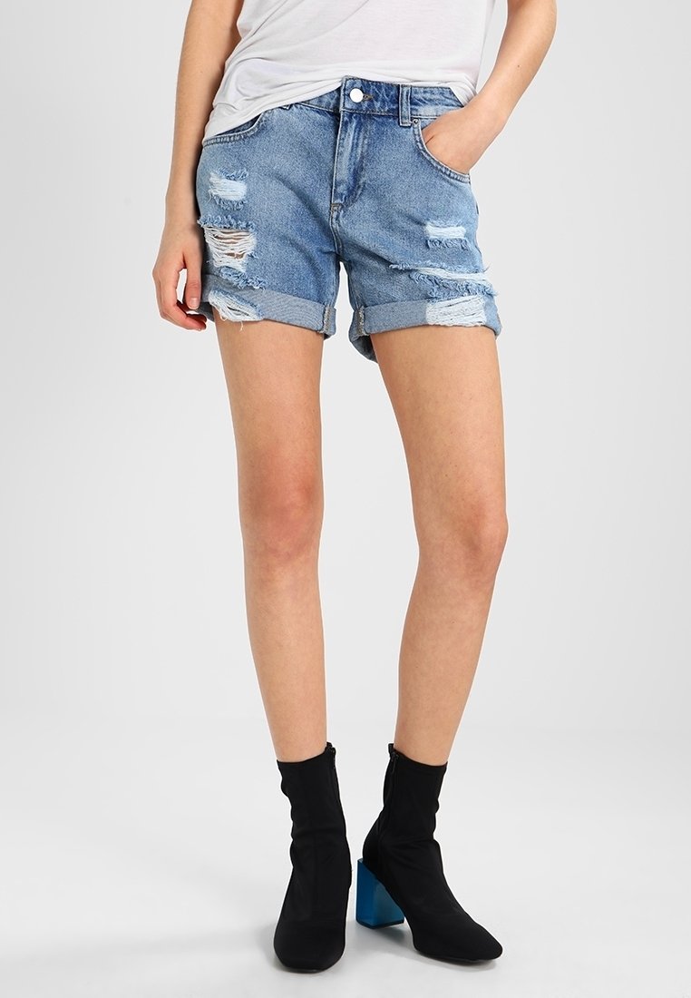 Shorts de mezclilla en azul claro con bordes deshilachados y detalles desgastados. Con puños doblados y un diseño clásico de cinco bolsillos. Combinados con botas negras hasta el tobillo.