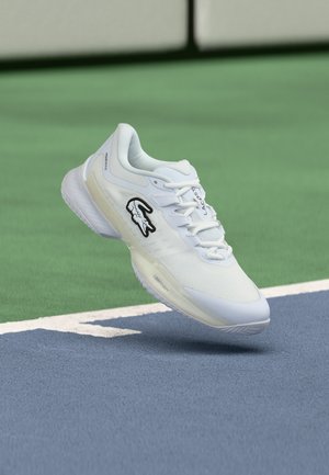 Chaussure de sport blanche Lacoste avec logo crocodile flottant au-dessus d'un court de tennis bleu et vert.