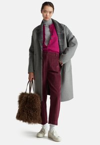 Šedý vlněný kabát přes růžový cardigan, šedý rolák a bordo manšestrové kalhoty; drží hnědou chlupatou tašku, má na sobě bílé tenisky.