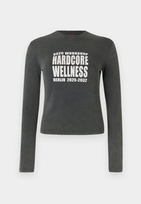 Maglietta a maniche lunghe di colore grigio scuro con testo bianco "032C Workshop Hardcore Wellness Berlino 2025-2032" centrato sul davanti.