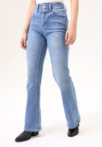 Jeans acampanados de cintura alta en azul claro con cierre de botones, dos bolsillos frontales y una sutil textura desgastada. Combinados con botas de tobillo negras de punta.