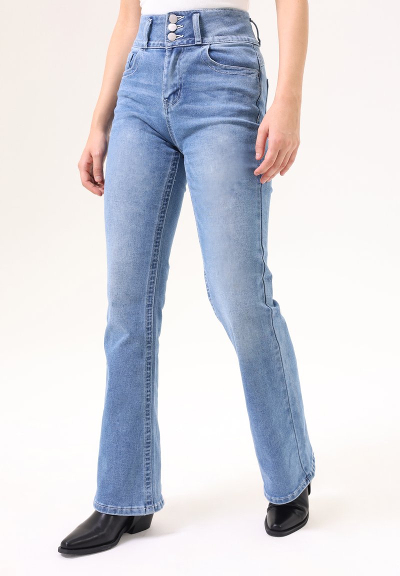 Jeans acampanados de cintura alta en azul claro con cierre de botones, dos bolsillos frontales y una sutil textura desgastada. Combinados con botas de tobillo negras de punta.