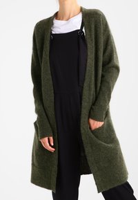 Cardigan long vert olive en tissu doux. Comprend un devant ouvert, des poignets côtelés et des poches latérales. Porté sur une tenue noire.