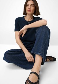 Navyblaues Baumwoll-T-Shirt, locker sitzende dunkelblaue Jeans und schwarze Netzschuhe. Einfaches Design mit minimalem Zubehör, sitzende Pose.