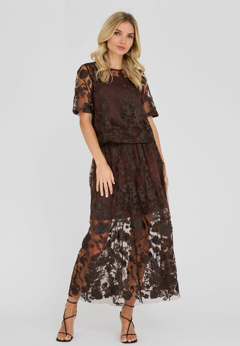 Femme portant une robe en dentelle florale noire transparente à manches courtes par-dessus une combinaison sombre, assortie à des sandales noires à talons hauts et à lanières.