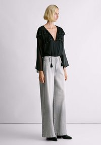 Kobieta ubrana w czarną przezroczystą bluzkę z falbankami i szare spodnie o szerokich nogawkach, stojąca na tle jasnego jednolitego tła, zwrócona bokiem.