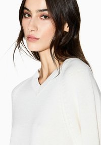 Weißer V-Ausschnitt-Pullover mit einer Strickstruktur, lockerem Sitz und dezenten diagonalen Nähten an den Schultern. Glattes, atmungsaktives Material.