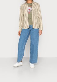 Kostkovaná béžová a zelená košile s dlouhým rukávem přes zelené tričko s grafikou, doplněná světlemodrými širokými džínami a bílými teniskami.