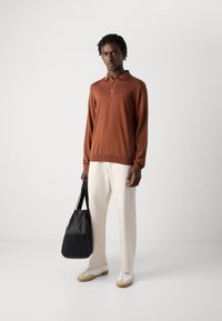 Mann in einem rostfarbenen Langarm-Polo, hellen Hosen und weißen Sneakern, der eine schwarze Tragetasche hält und vor einem schlichten weißen Hintergrund steht.