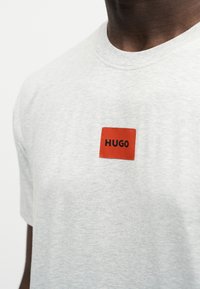 Gros plan d'un T-shirt gris clair avec un petit patch carré rouge centré sur la poitrine, avec le mot « HUGO » en lettres noires.
