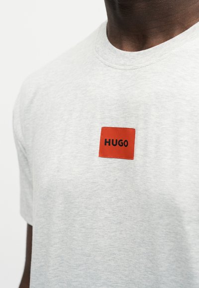Gros plan d'un T-shirt gris clair avec un petit patch carré rouge centré sur la poitrine, avec le mot « HUGO » en lettres noires.