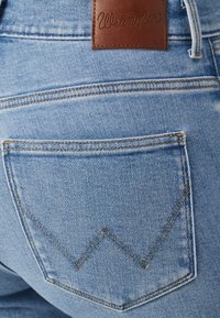 Ljusblå denimjeans med en bakficka som har ett zickzackstygnsmönster och ett brunt lädermidjeband med präglad texten "Wrangler."