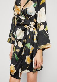 Robe enveloppante en satin noir à motif floral, avec des manches longues et une taille nouée, arborant des roses claires et des feuilles vertes sur un fond sombre.