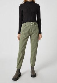 Top de manga larga negro de canalé combinado con pantalones de tiro alto de color verde claro, con una textura suave y cierre de botón frontal. Botines negros.