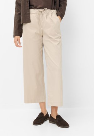 Beige bukser med vidde ben og snøre i taljen, båret med mørkebrune loafers og en mørk top, vist på en stående person mod hvid baggrund.