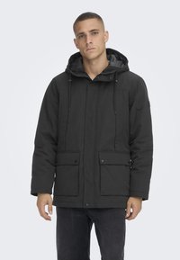 Parka negra aislada con capucha, que cuenta con dos bolsillos frontales, cierre de cremallera en la parte delantera y detalles de cordón para un ajuste ajustable. Textura suave.