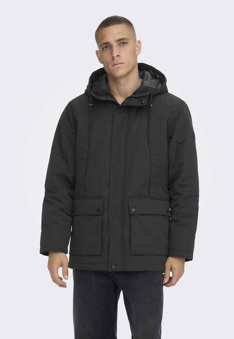 Parka negra aislada con capucha, que cuenta con dos bolsillos frontales, cierre de cremallera en la parte delantera y detalles de cordón para un ajuste ajustable. Textura suave.