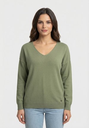 Mujer de cabello largo y oscuro, vistiendo un suéter verde con cuello en V y jeans azul claro, de pie frente a un fondo blanco liso.