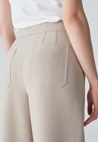 Pantalons beiges ajustés avec une texture lisse, jambes larges, et poches plaquées doubles à l'arrière. Des coutures contrastées soulignent les poches.
