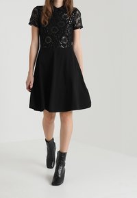 Svart klänning med blommig spetsöverkvlpning, korta ärmar och en utsvängd kjol. Tillsammans med svarta ankelstövlar. Enkelt mönster med texturerade kontraster.