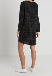 Nainen, jolla on päällään musta pitkähihaisen mini-mekko, jossa on pieniä vaaleanpunaisia pisteitä, yhdistettynä valkoisiin tennareihin, seisoo yksinkertaista vaaleaa taustaa vasten.