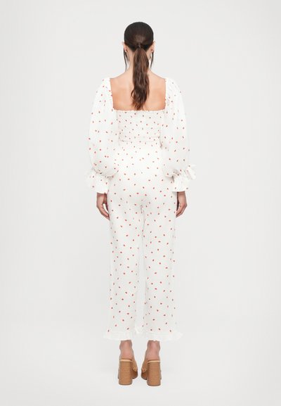 Hvid jumpsuit med røde prikker, smocket liv og puffede ærmer. Flared ben og flæse detalje i kanten med åben ryg. Brune blokhæle.