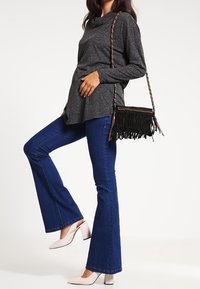 Grauer Cowl-Neck-Top, dunkelblaue Schlagjeans, weiße spitze Absätze und eine schwarze Fransen-Crossbody-Tasche.