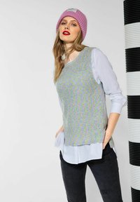 Un maglione lavorato a maglia in filato multicolore è posato su una camicia azzurra. La modella indossa un berretto rosa; jeans neri strappati con tasche visibili.