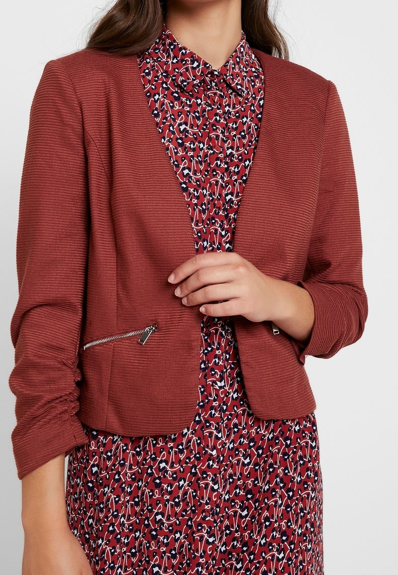 Blazer en velours rouge rouille avec une poche zippée sur le côté. Manches longues avec des poignets froncés, porté par-dessus une robe à motif floral.