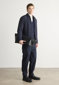 Chaqueta de botonadura navy con dos bolsillos en el pecho, superpuesta a un suéter texturizado navy. Bolso de mano negro alargado, pantalones navy ajustados y botas negras.